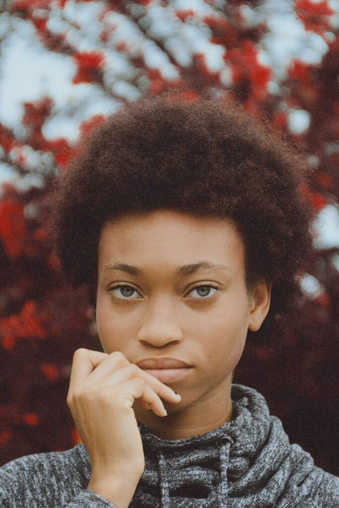 Portrait of a young woman with natural afro-textured hair, wearing a gray hoodie, posing thoughtfully with her hand near her face. The vibrant red foliage in the background adds contrast to her calm demeanor. Professional CV writing services