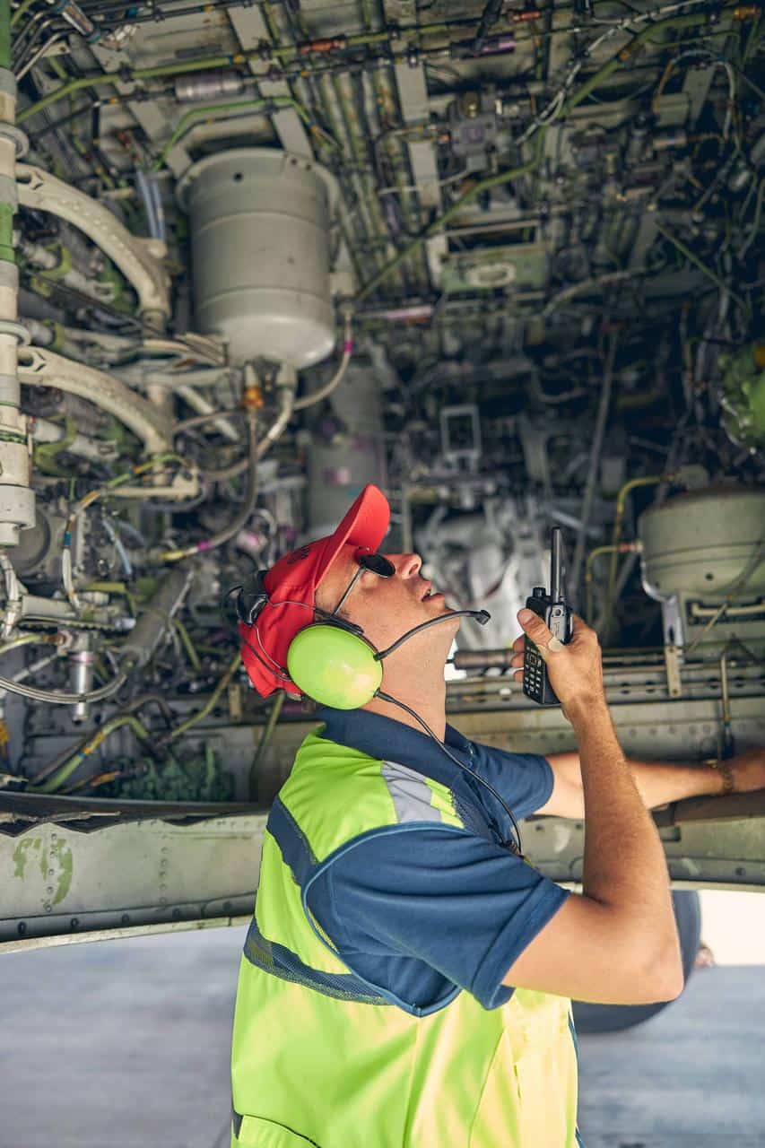 Aircraft mechanic inspecting airplane engine systems with headset and radio for maintenance operations.