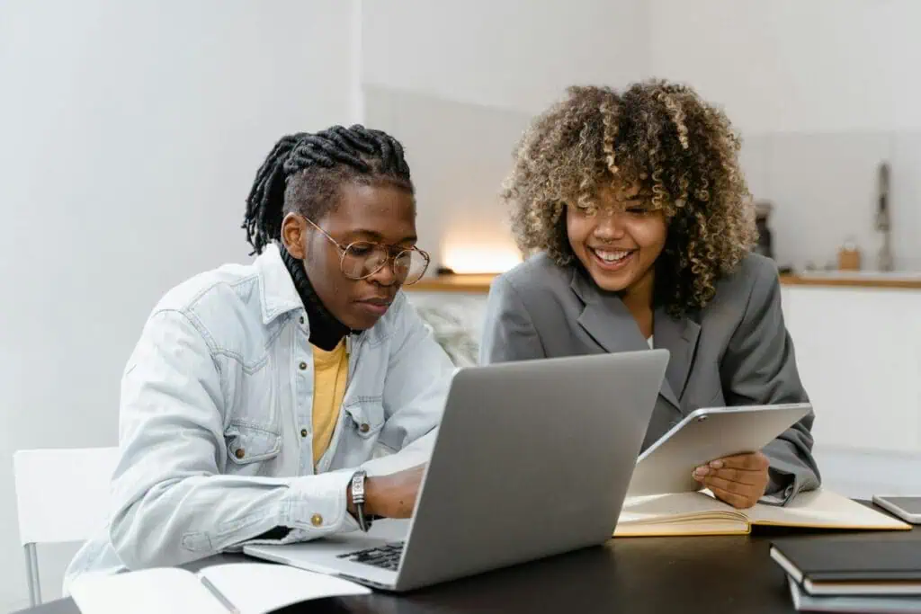 Two Algerian students collaborating at a desk, using a laptop and a tablet in a bright modern setting.