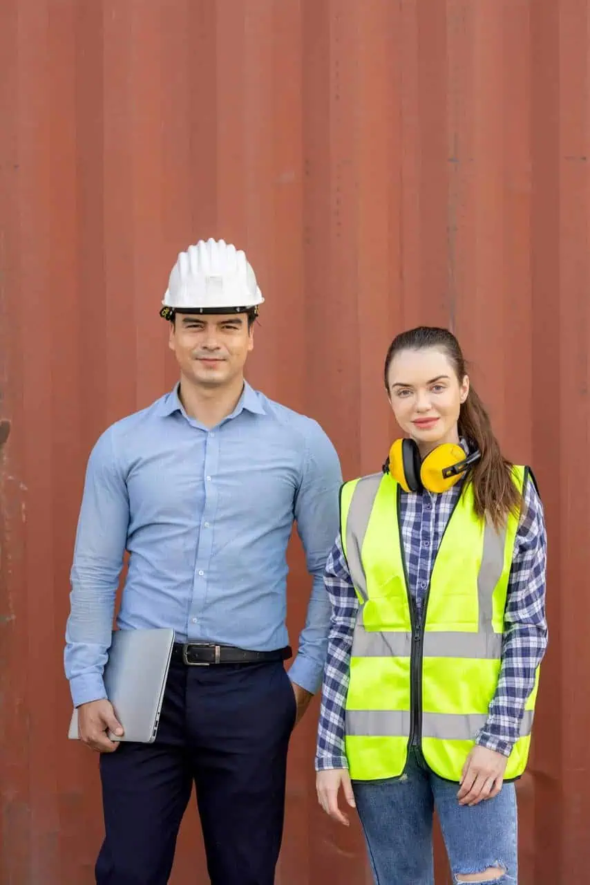 A man in a white safety helmet and blue shirt stands beside a woman in a reflective vest and earmuffs, both posing confidently in front of a red industrial container.