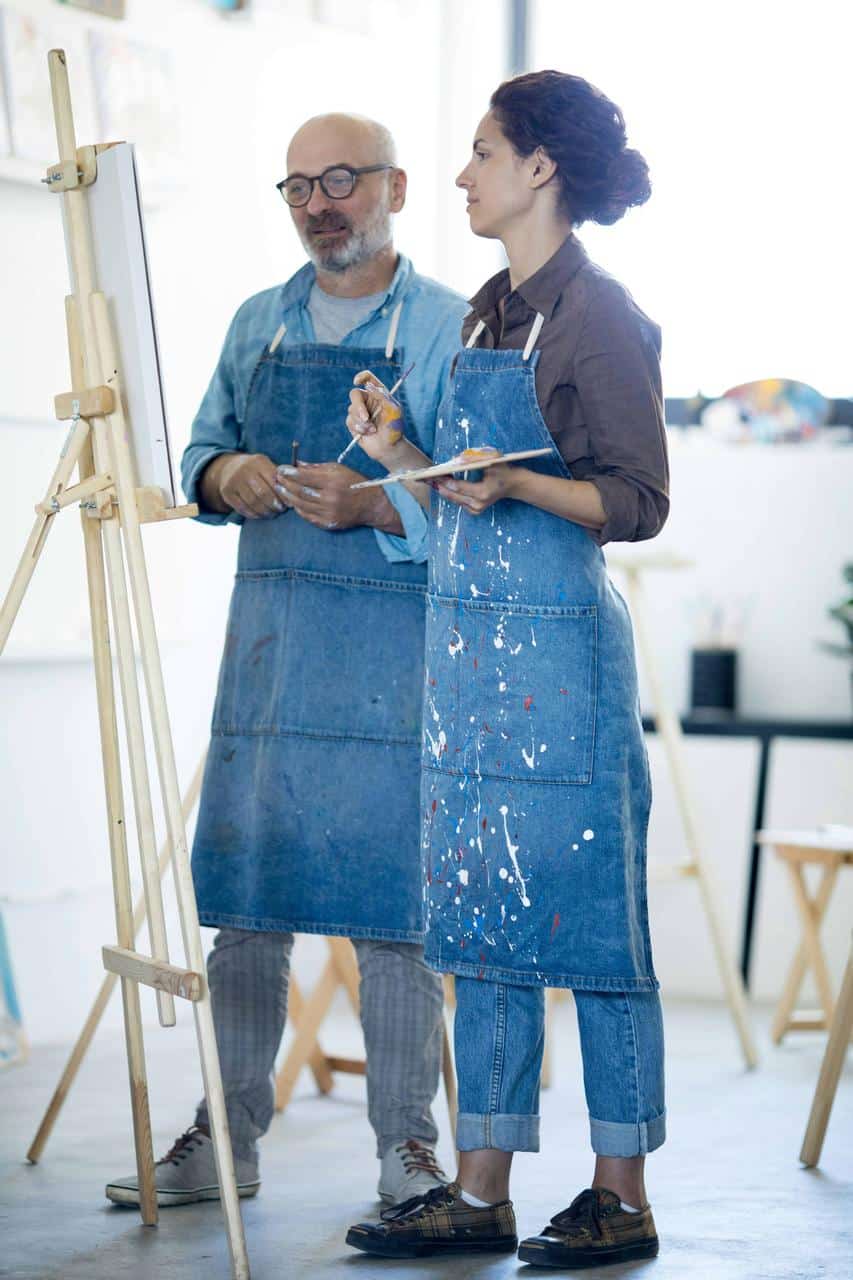 Art teacher and student wearing denim aprons stand beside an easel, holding paintbrushes and a palette during an art lesson.