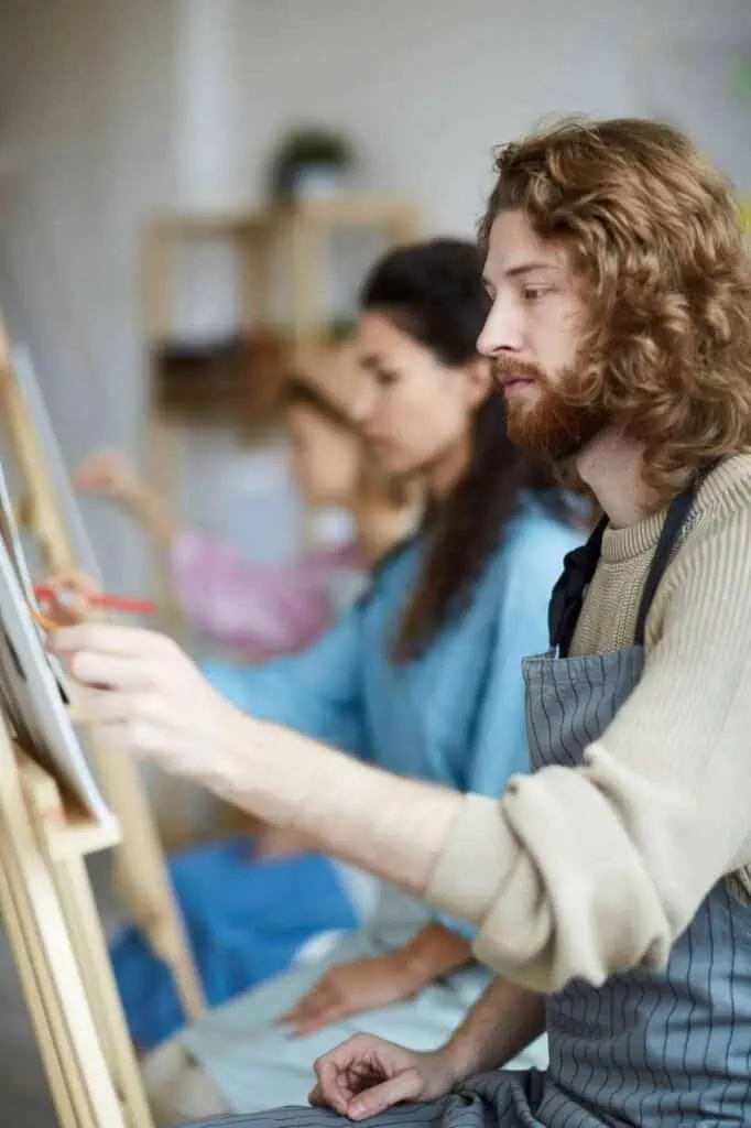 A man wearing a striped apron concentrates on his artwork during an arts and crafts workshop, surrounded by other focused participants.