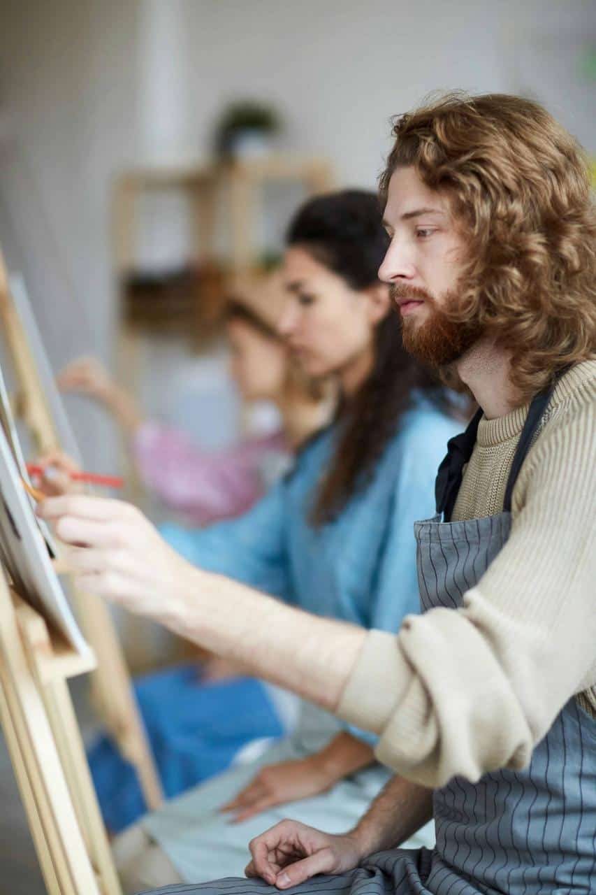 A man wearing a striped apron concentrates on his artwork during an arts and crafts workshop, surrounded by other focused participants.
