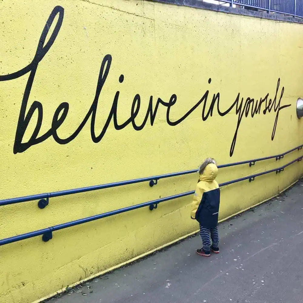 Child in a yellow jacket looking at an inspiring 'Believe in Yourself' mural on a wall.