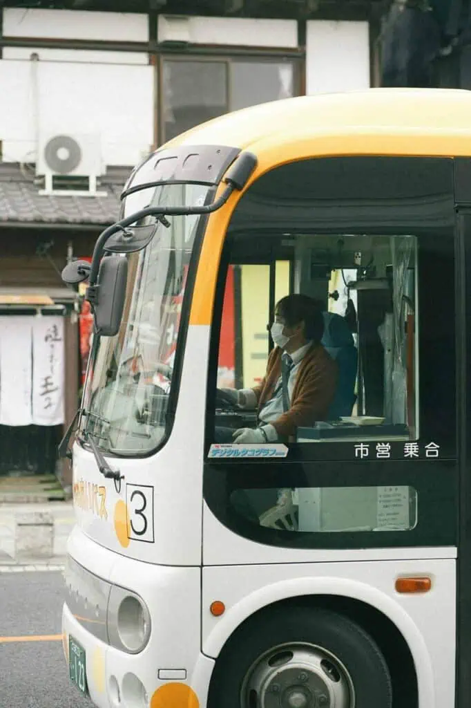 Professional bus driver wearing a mask while driving a public transport vehicle in an urban setting.