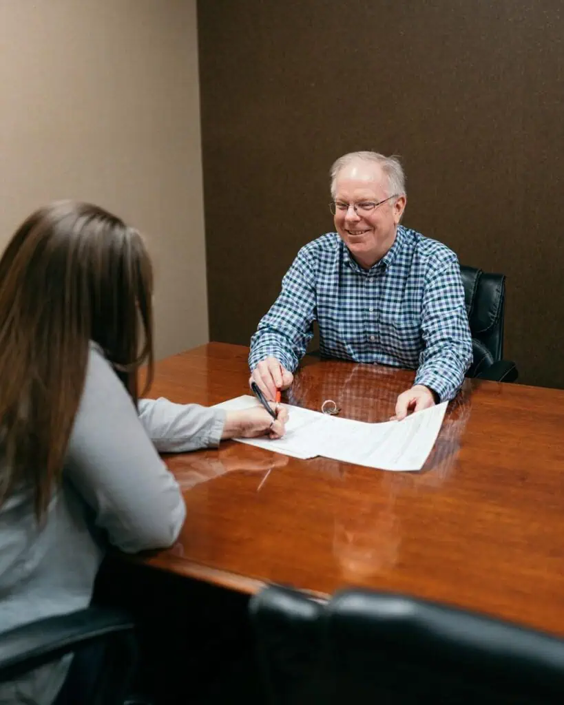 An older male career advisor in a checked shirt smiles while guiding a young woman through a document during a one-on-one consultation at a wooden office desk.