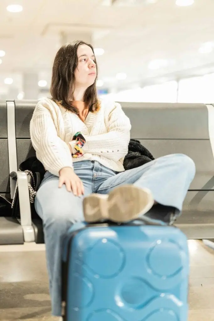 Young Chilean woman sitting at an airport with her legs propped on a blue suitcase, wearing a cozy sweater and jeans, looking off into the distance
