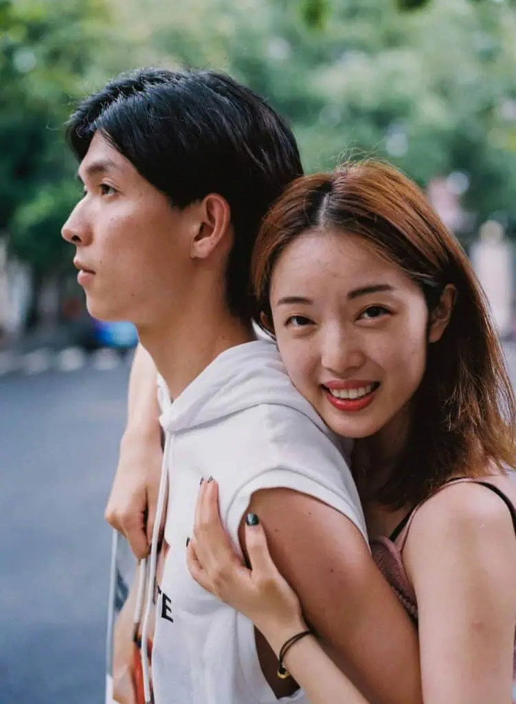 Close-up of a young student couple outdoors, with a smiling woman hugging her partner from behind as he looks away thoughtfully.