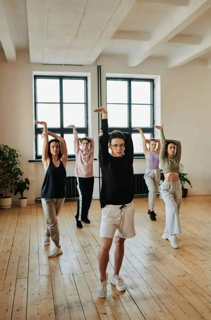A dance instructor leads a group of students through a synchronized routine in a bright studio with wooden floors and large windows.