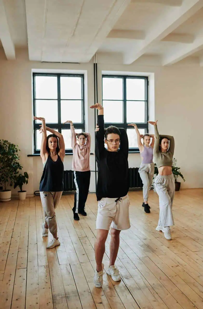 A dance instructor leads a group of students through a synchronized routine in a bright studio with wooden floors and large windows.