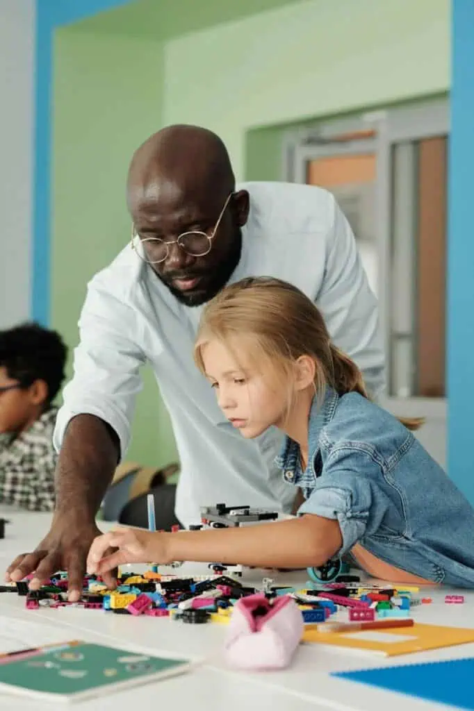 Male preschool teacher helps a young girl build a model using colorful plastic blocks at a classroom table.