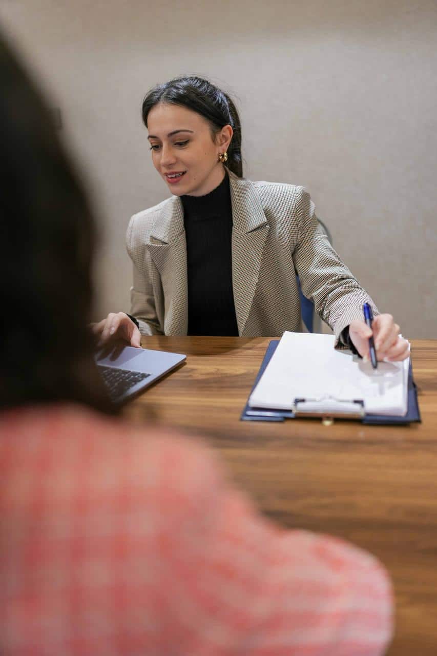 A young woman in a business blazer takes notes on a clipboard while attentively assisting a client at a wooden office desk.