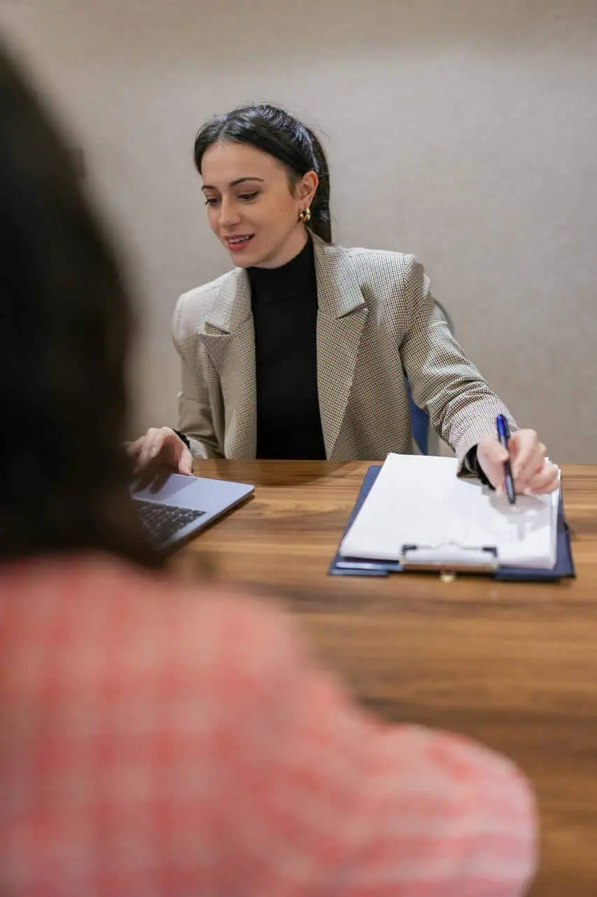 A young woman in a business blazer takes notes on a clipboard while attentively assisting a client at a wooden office desk.