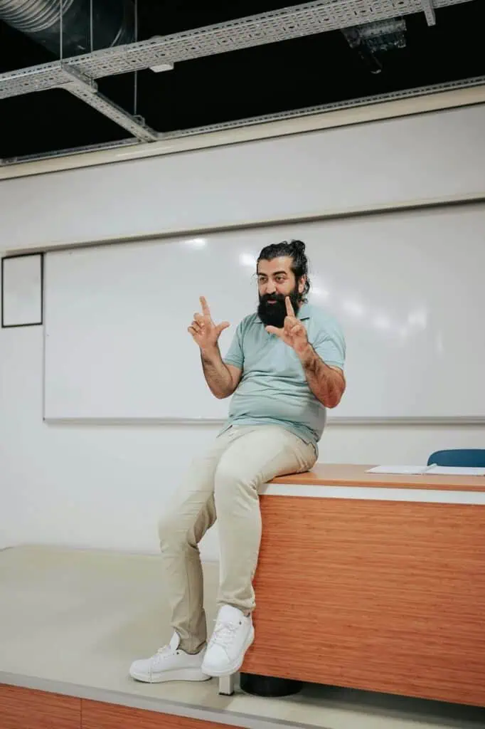 University professor in casual attire sits on a desk in front of a whiteboard, using hand gestures during a lecture.