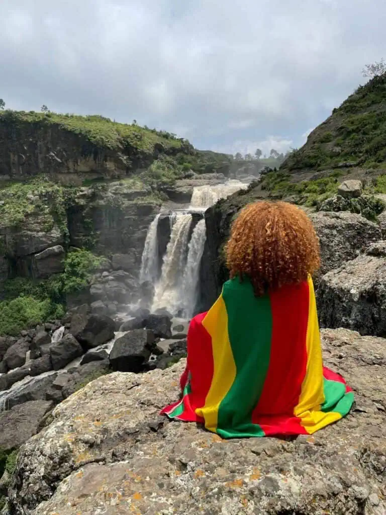 Student wrapped in the Ethiopian flag overlooking a scenic waterfall, symbolizing education and cultural pride.