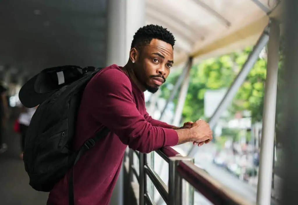 Ethiopian graduate in a burgundy sweater and black backpack leaning on a railing, looking thoughtfully into the distance in an outdoor urban setting.