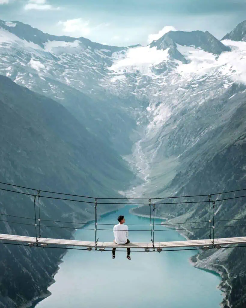 European student with a Professional Student CV sitting on a suspension bridge, overlooking a breathtaking alpine valley with snow-capped mountains and a glacial lake.