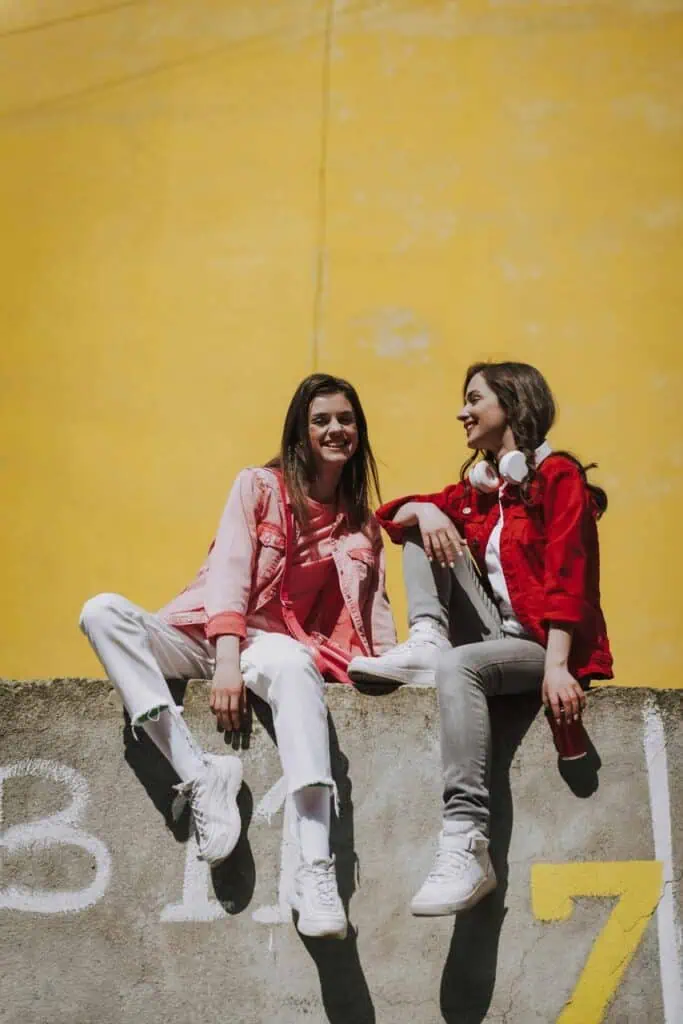 Two young German students sitting on a concrete wall against a bright yellow background, smiling and talking about their career options.