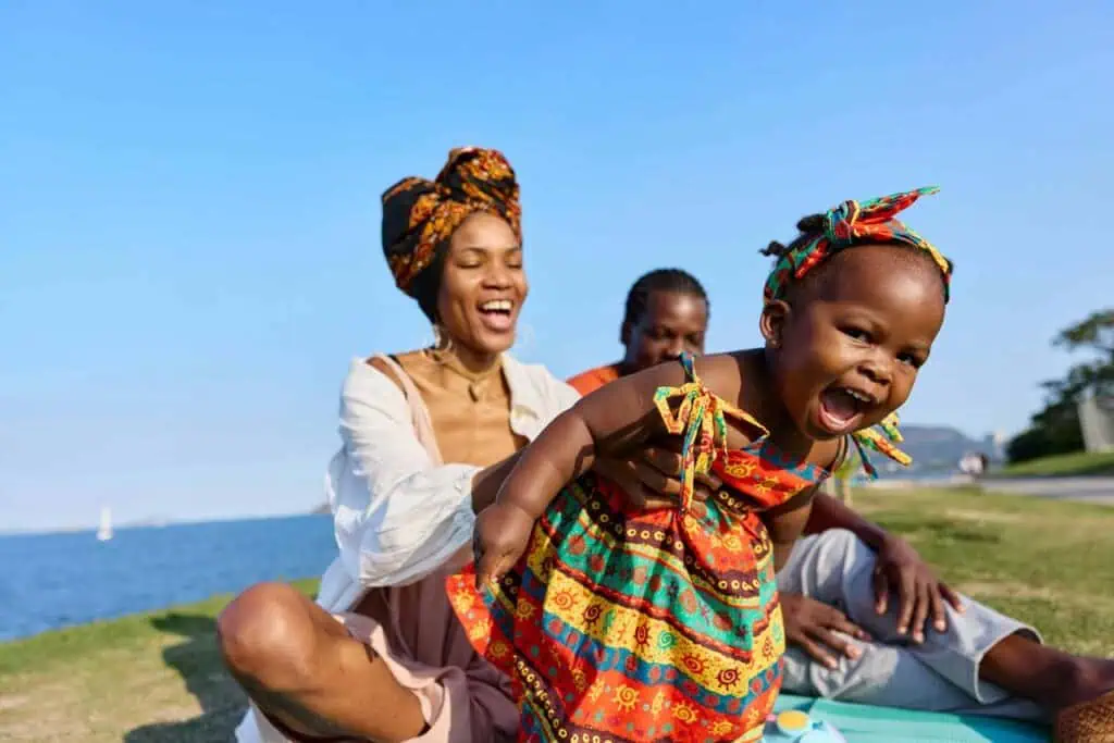 A Ghanaian family sitting by a waterfront, sharing a joyful moment. A young child dressed in vibrant traditional Ghanaian clothing is playfully held by a smiling woman wearing a matching headwrap, as they enjoy the sunny outdoors.
