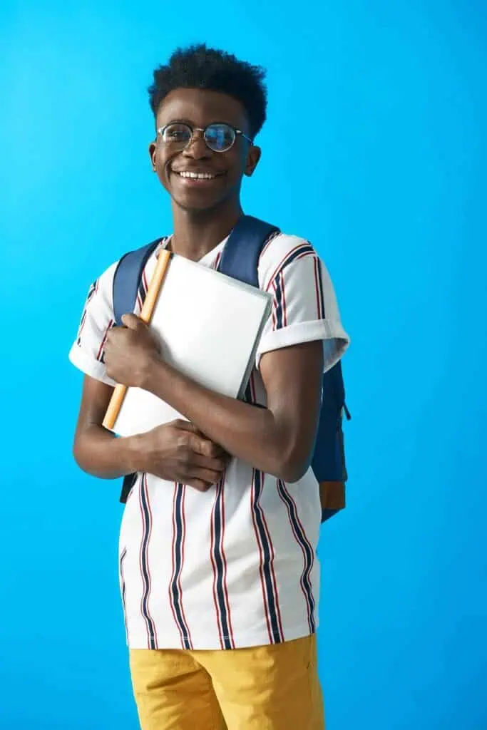 A young Ghanaian student wearing glasses and carrying a backpack, holding books against a bright blue background, smiling confidently as he considers his future career options.