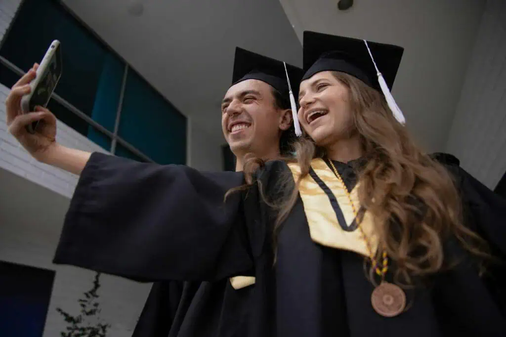 Two graduates in black caps and gowns taking a joyful selfie outdoors, celebrating their academic achievement.