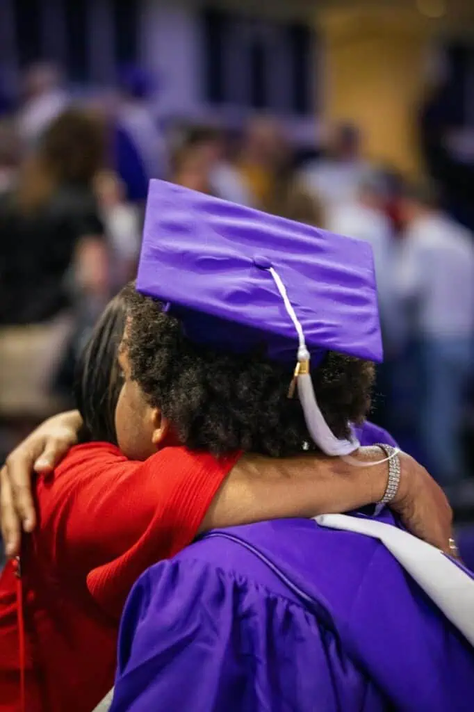 Graduate in a purple cap and gown hugs a loved one in celebration, surrounded by a blurred crowd at a graduation ceremony.