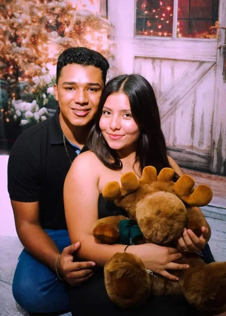 A young Guatemalan couple posing in front of a festive holiday background, with the woman holding a large plush moose toy. They share a moment of closeness while reflecting on their immigration journey.