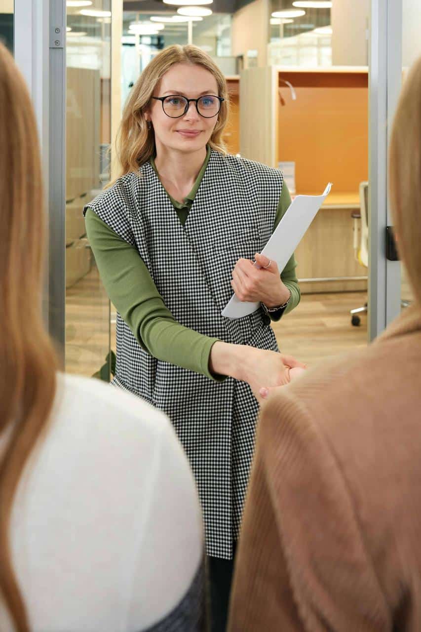 A smiling HR professional wearing glasses and holding a clipboard warmly greets two job candidates inside a modern office environment.