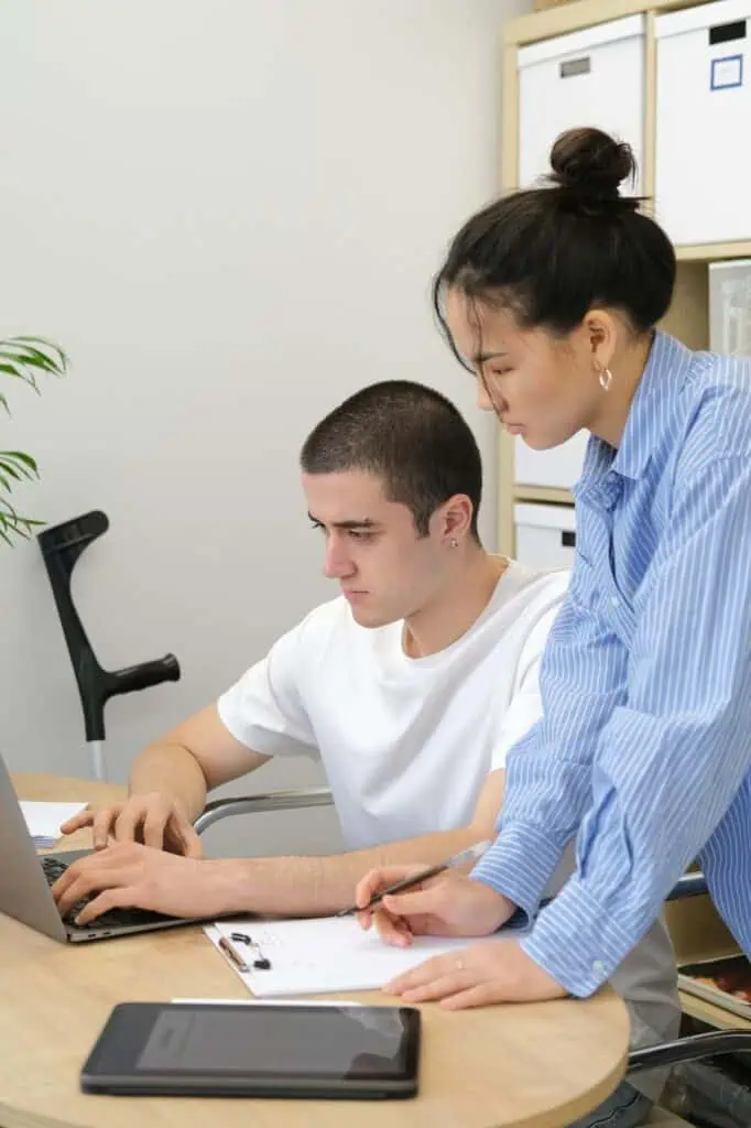 Young man in a wheelchair receives hands-on tutoring from a woman while working on a laptop, with study materials on the desk.