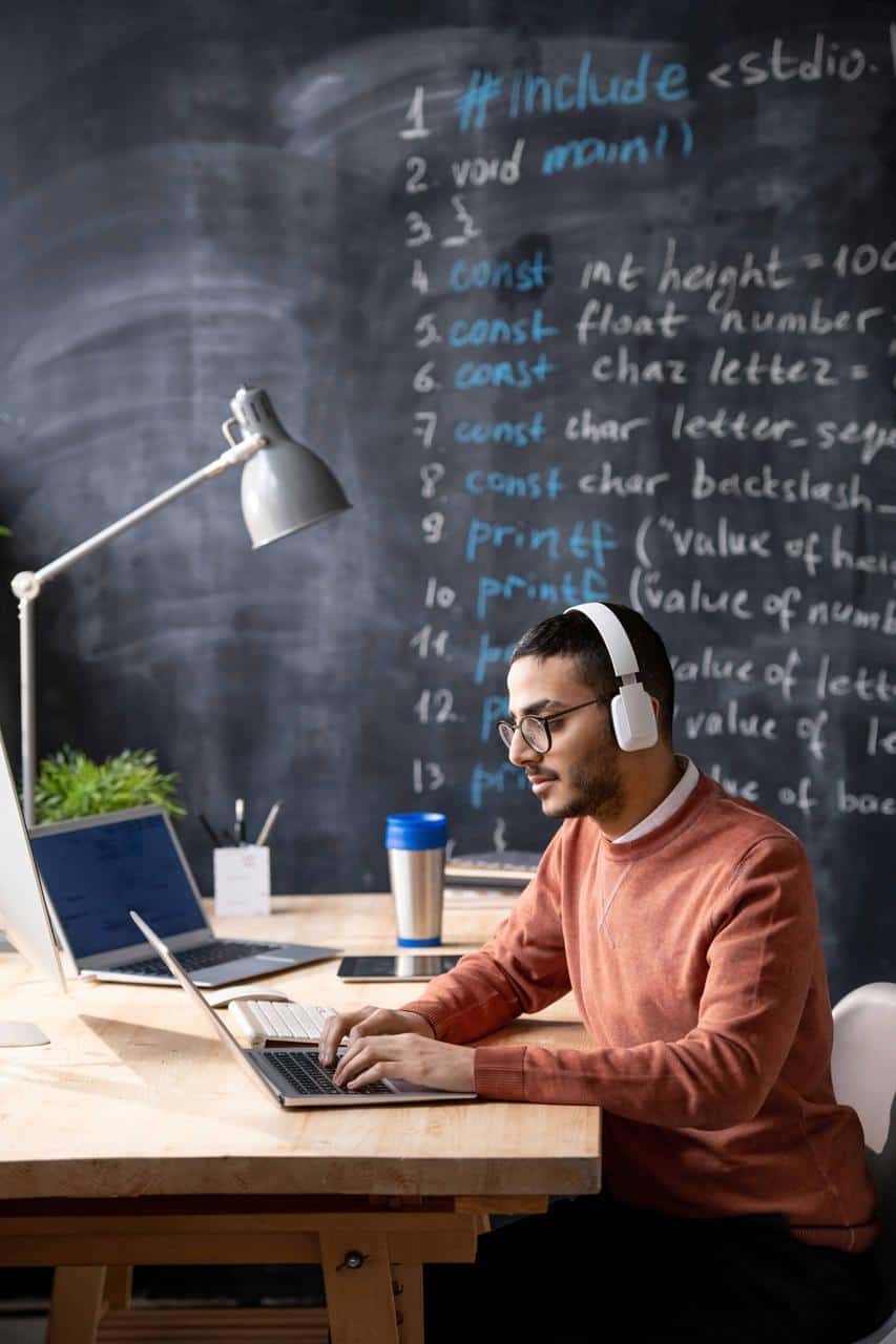 Male IT support specialist wearing headphones and working on a laptop in a modern classroom with coding written on a chalkboard behind him.