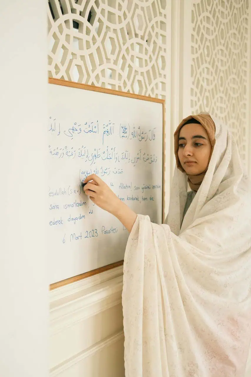 Female language tutor in a traditional outfit teaching Arabic on a whiteboard with script and translation.