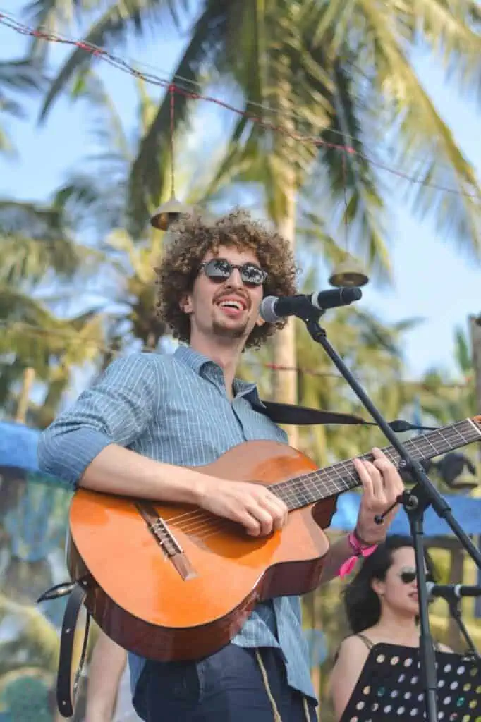 A smiling male musician plays an acoustic guitar and sings into a microphone on an outdoor stage, with palm trees in the background.