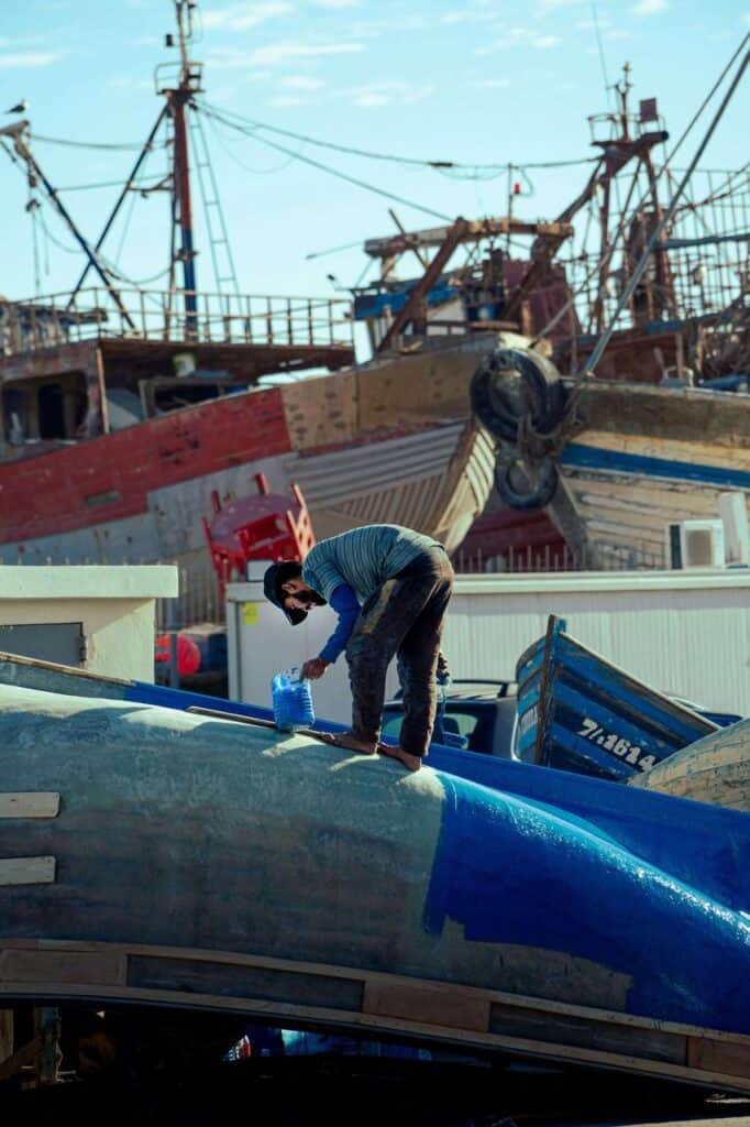 Longshoreman painting fishing boat in shipyard with docked vessels in background