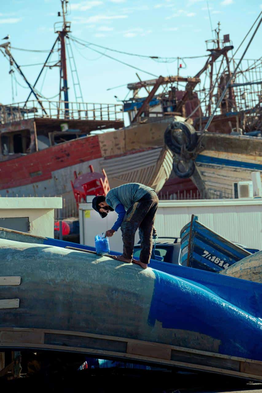 Longshoreman painting fishing boat in shipyard with docked vessels in background