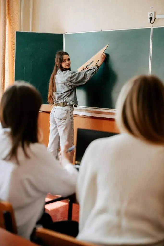 Female student in a denim outfit uses a large triangle ruler to draw on a green chalkboard while teaching classmates in a classroom.