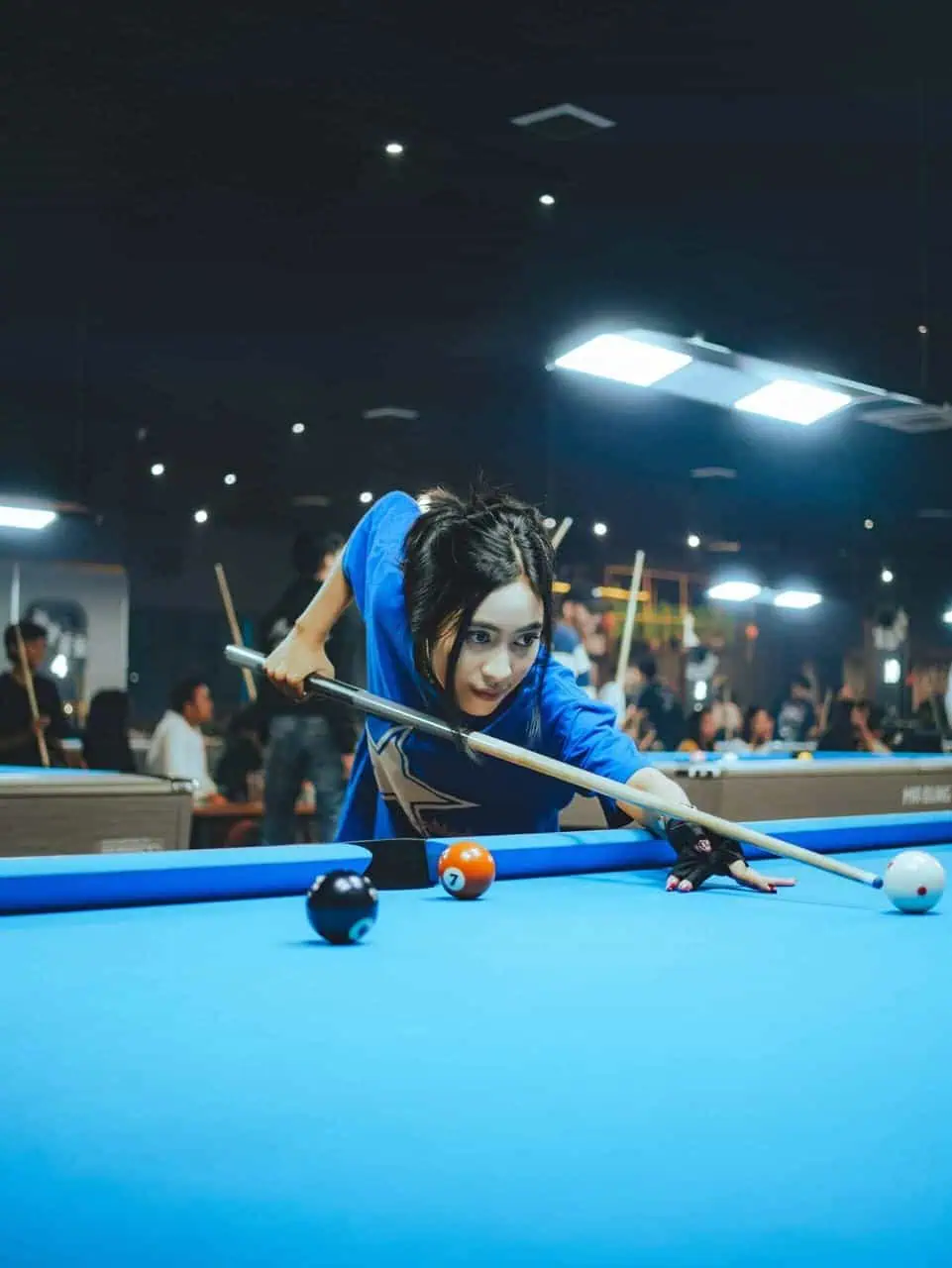 A focused young woman lines up a shot at a billiards table in a vibrant and busy social club environment.