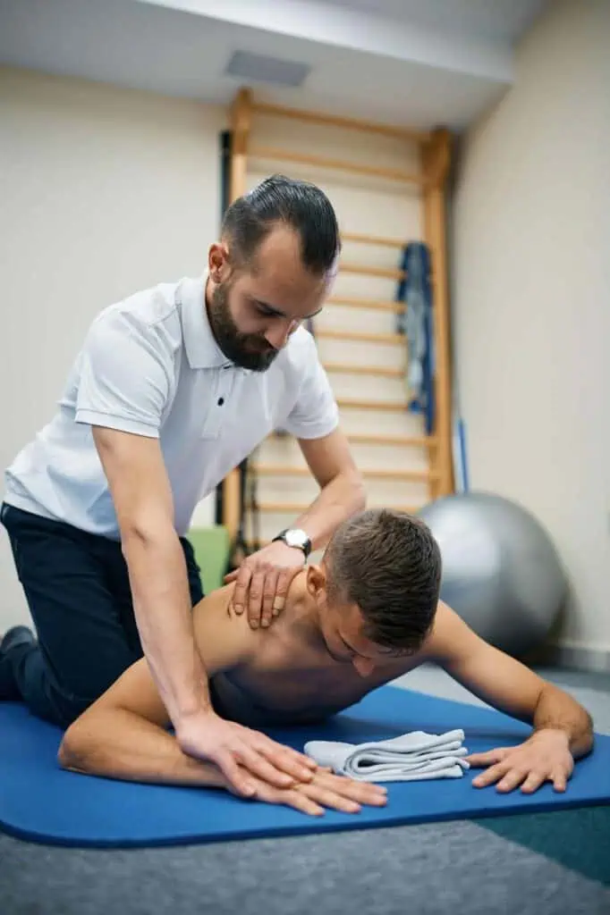 A sports physiotherapist in a white polo shirt performs a manual therapy technique on a male athlete lying on a mat during a rehabilitation session.