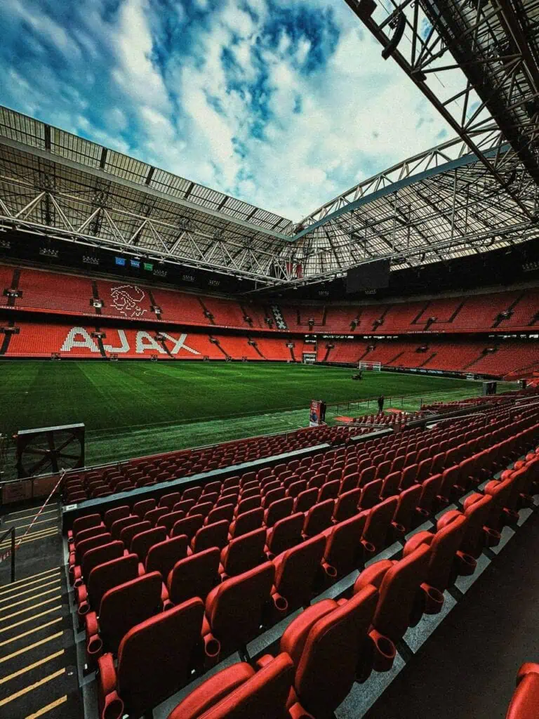 Wide-angle view of an empty professional football stadium with red seats, green pitch, and a partially open retractable roof under a cloudy sky.