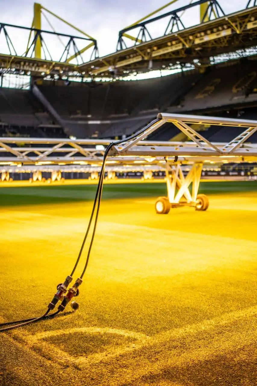 Close-up of turf lighting equipment on a sports stadium field, with cables attached and yellow grow lights illuminating the grass.