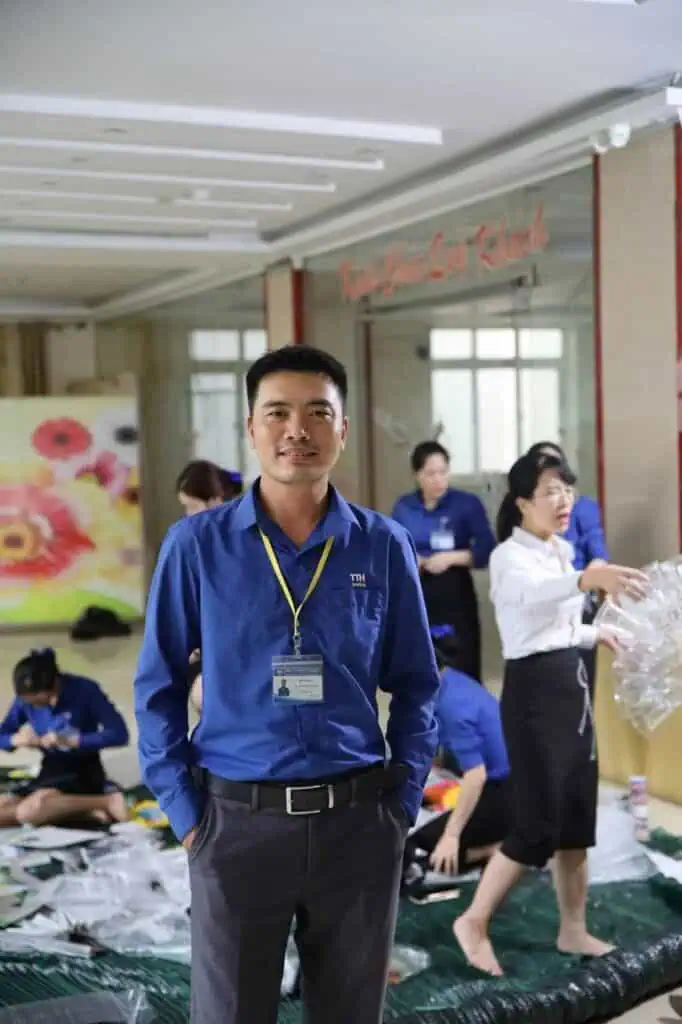 A student affairs coordinator in a blue shirt and name badge stands smiling in a student activity space, with students and staff engaging in a background project.