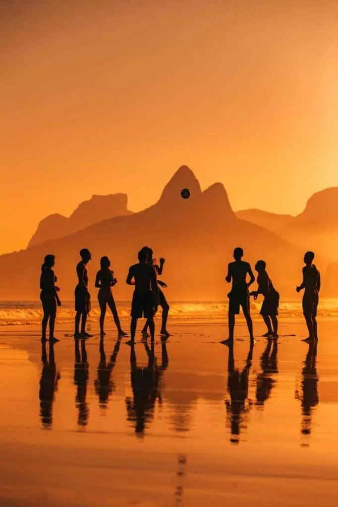 Silhouette of a group of students playing soccer on the beach at sunset with mountains in the background, reflecting golden hues on the sand.