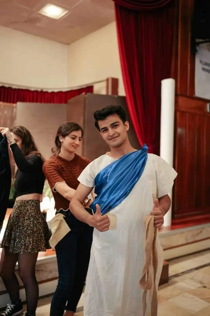 A young man in a toga costume smiles and gives a thumbs up backstage, while others help with costumes before a theatrical performance.