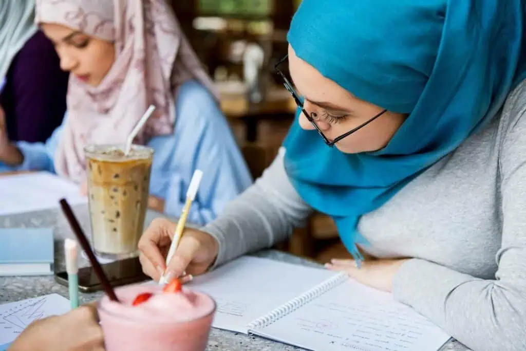 Two young Egyptian student in hijabs studying together at a café table with notebooks, iced coffee, and a strawberry smoothie.