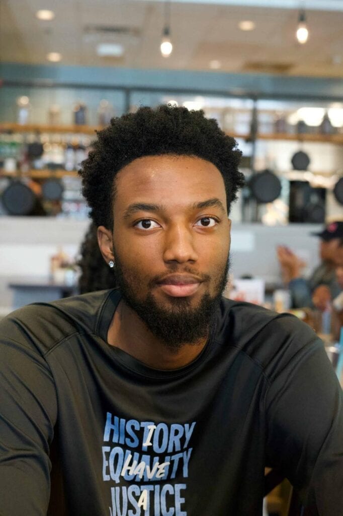 Young Columbian student with a beard and curly hair sitting in a cafe, wearing a black shirt with the words 'History, Equality, Justice,' looking thoughtfully at the camera.