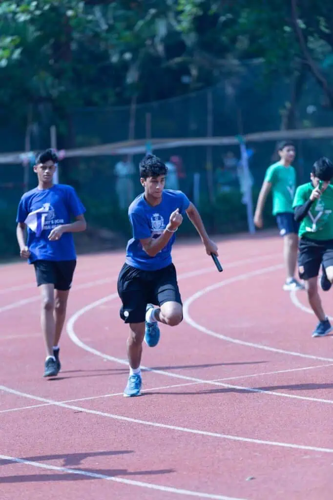 Boys in team uniforms compete in a relay race on a red running track during a school athletics event, each holding a baton mid-sprint.