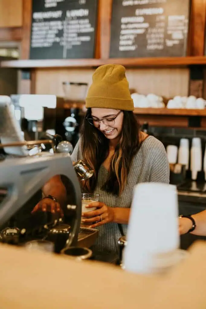 Smiling barista preparing a coffee behind the counter in a trendy café, showcasing hospitality and customer service skills for student job seekers.