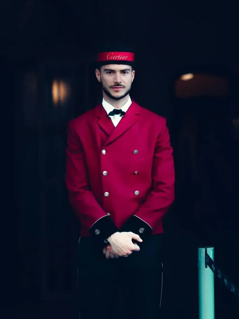 Hotel bellhop in formal red uniform standing at an entrance—representing front-of-house hospitality and entry-level service roles.
