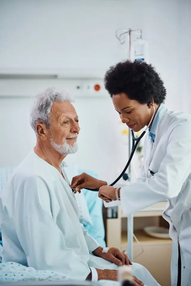 Female cardiologist examining an elderly male patient in a hospital room using a stethoscope.