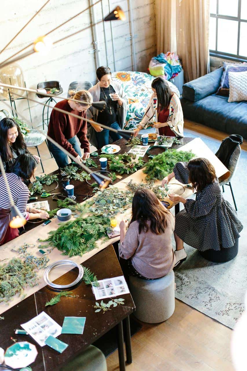 A diverse group of people collaborating around a large table, engaged in a creative community activity led by a program facilitator in a cozy, inclusive space.