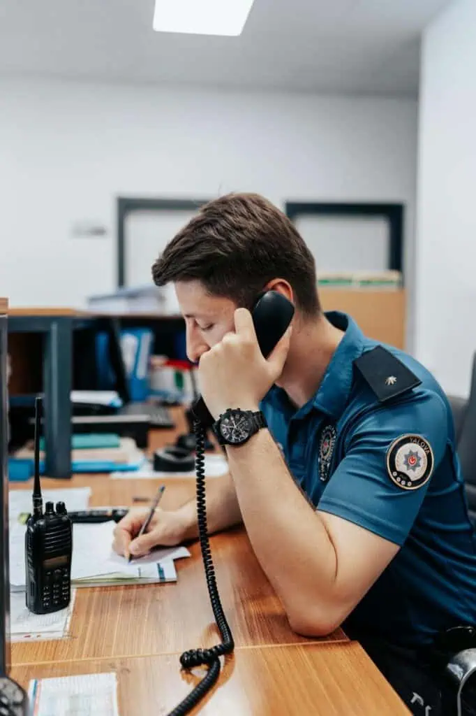 A government call center operator in uniform answering a phone and recording information at a workstation equipped with documents and a radio.